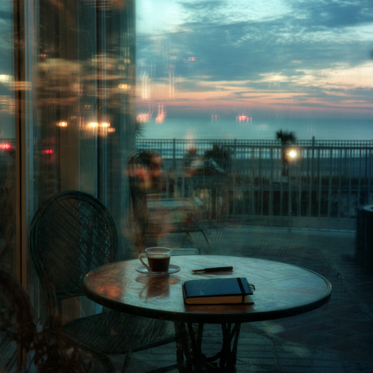 dawn veranda at a seaside hotel: a single coffee cup cooling on a small table; distant police lights reflected on the glass door; a closed black notebook beside the cup