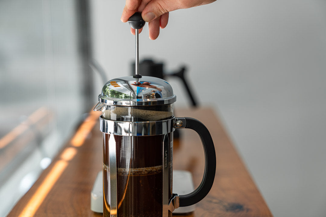 hands pressing down a French press with rich coffee and rising steam in soft natural light