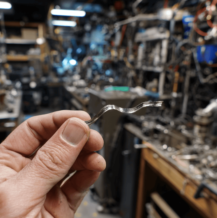 Self-bending metallic strip curling upward in response to human touch, representing the mysterious low-threshold alloy.