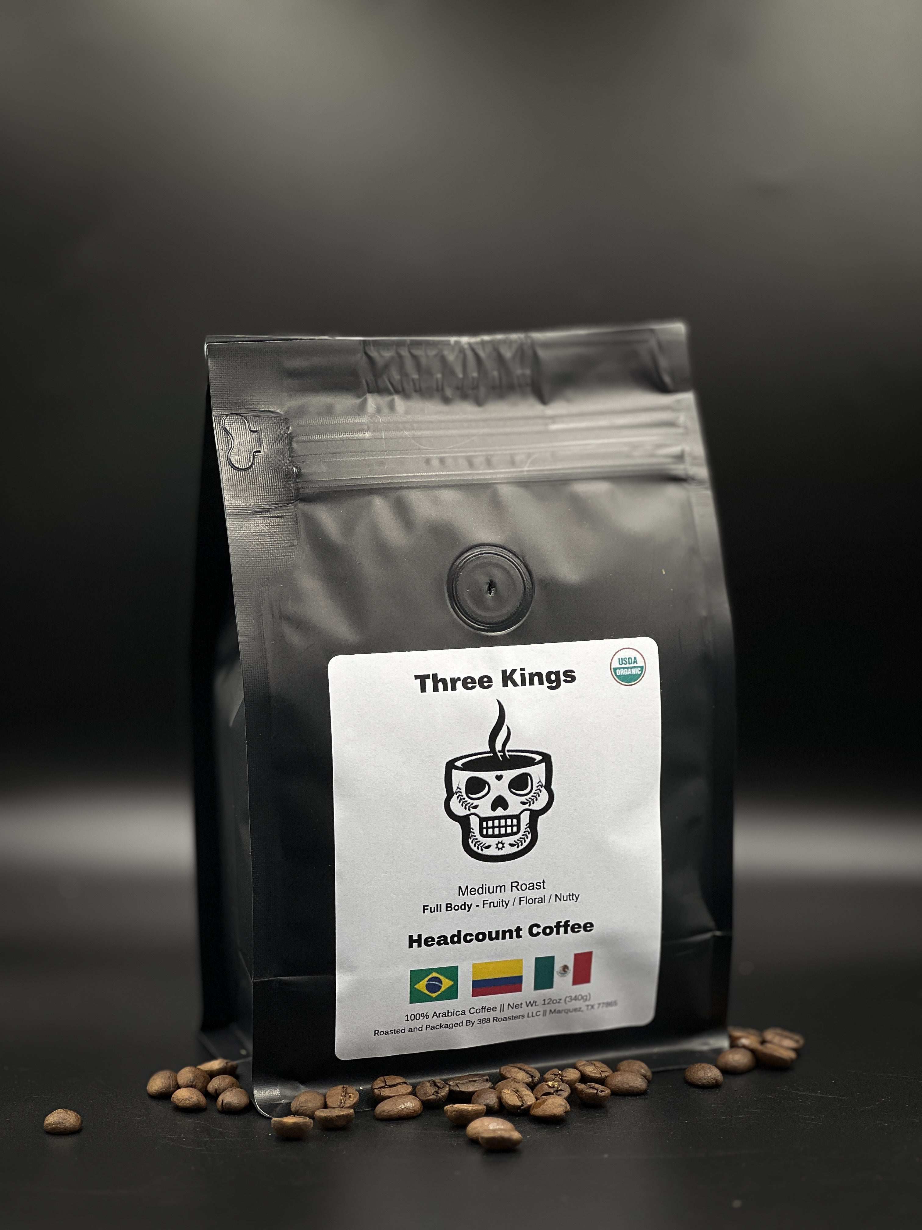 A package of Headcount Coffee branded coffee with the name 'Three Kings' displayed on the label. The package is black with a graphic of a skull and crossbones, and it is placed against a dark background with coffee beans scattered around it.